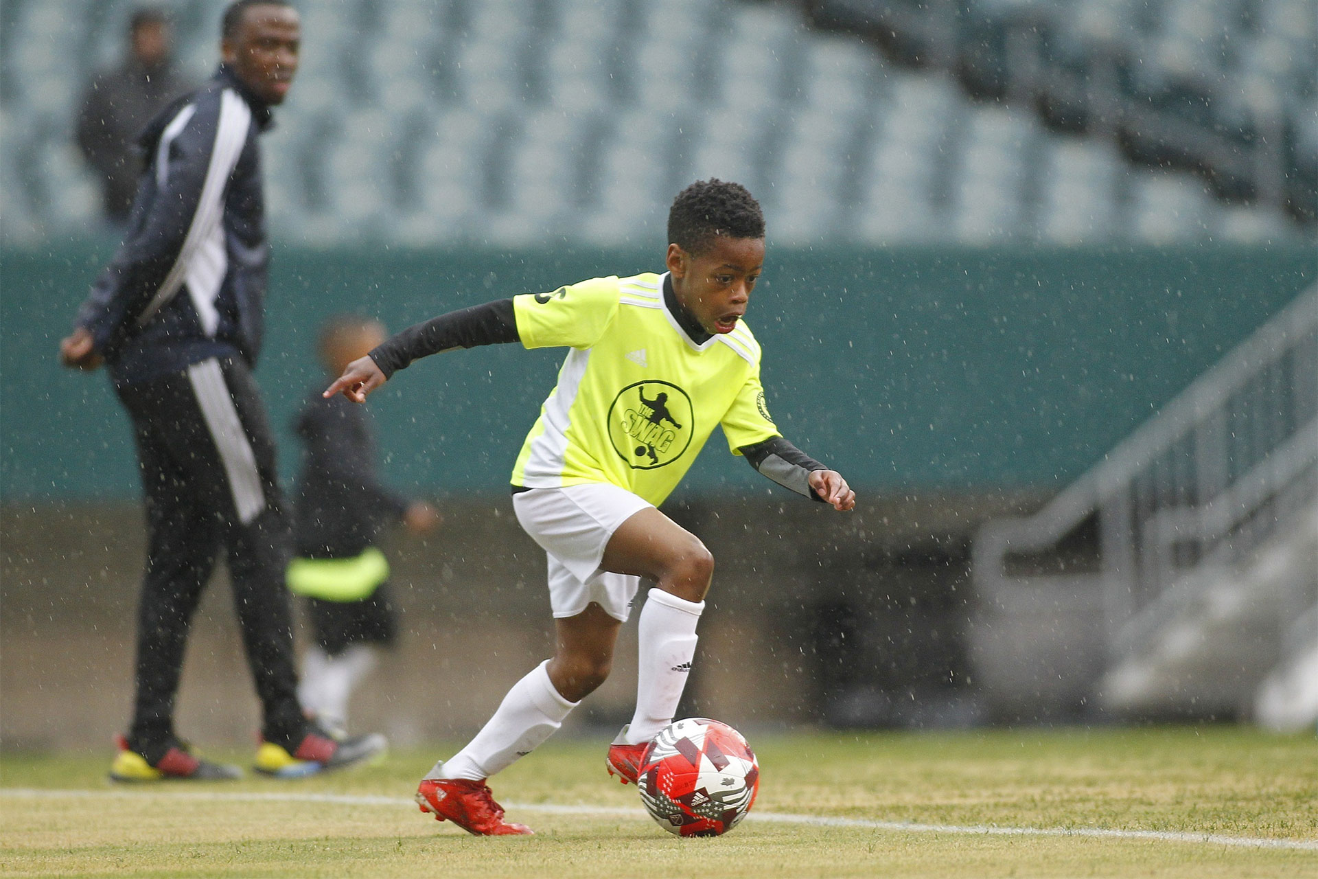A child playing soccer wearing a jersey labeled, "The Swag." A coach looks on from the background.